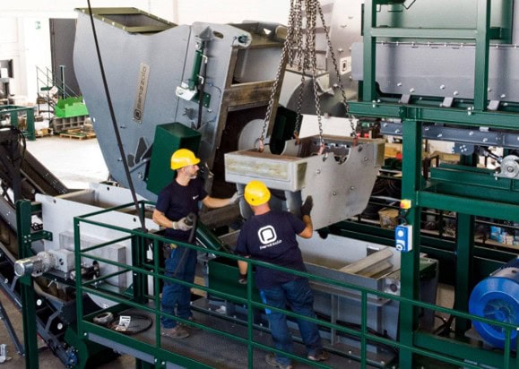 Workers operating a large industrial hammer mill for waste processing.